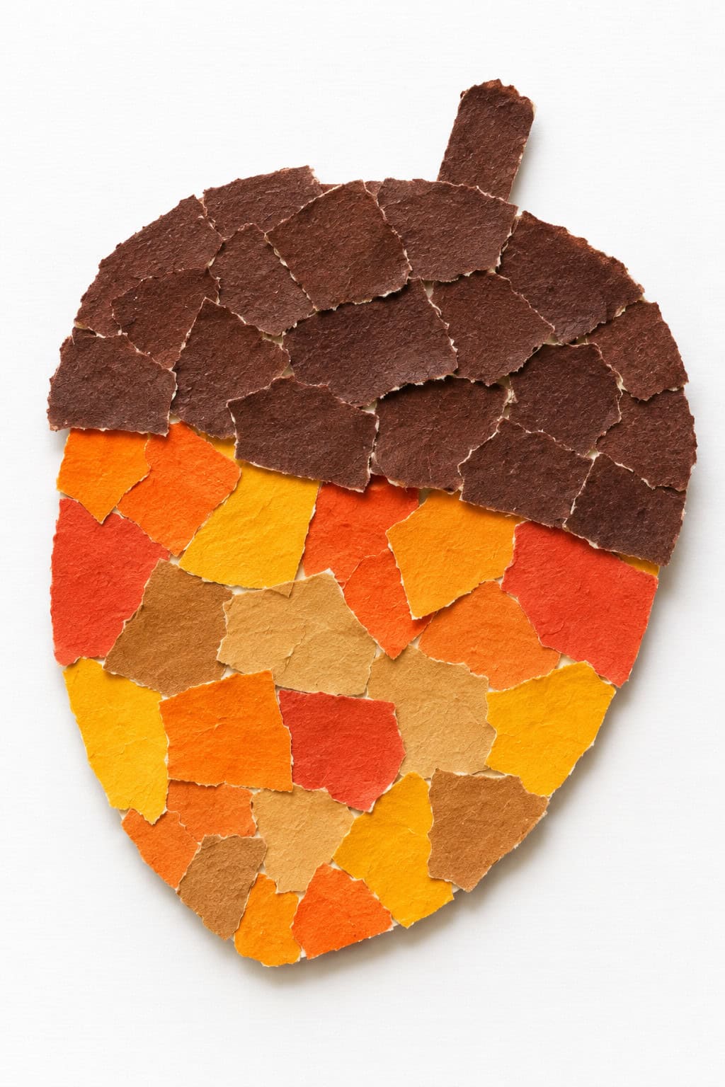 Acorn-shaped collage on a white background made from torn paper pieces. The rounded lower section uses orange, yellow, tan, and red scraps, while the cap and short stem are covered with overlapping dark brown pieces.