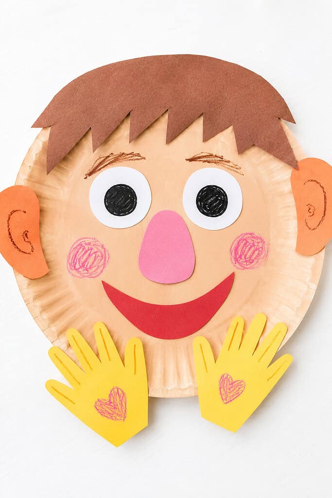 A paper plate face with brown paper hair, large white-and-black eyes, orange ears, a pink nose, red smile, rosy cheeks, and two yellow paper hands on a bright white table.