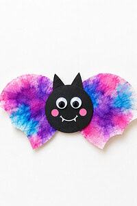 A handmade paper bat with a round black face, pointed ears, googly eyes, pink cheeks, a white smile, and wide pink, purple, and blue watercolor wings on a bright white background.