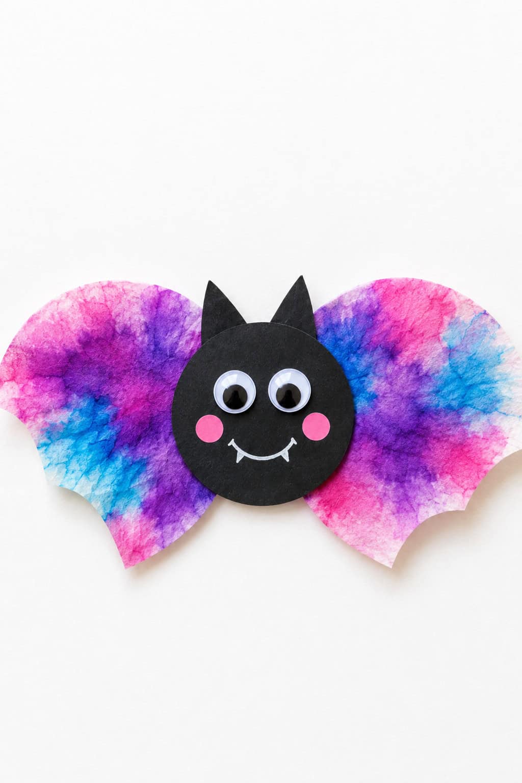 A handmade paper bat with a round black face, pointed ears, googly eyes, pink cheeks, a white smile, and wide pink, purple, and blue watercolor wings on a bright white background.