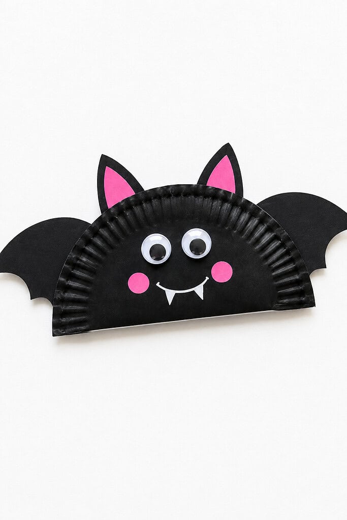 A black paper plate bat with wide paper wings, pink inner ears and cheeks, googly eyes, a white smile, and two small fangs on a bright white background.