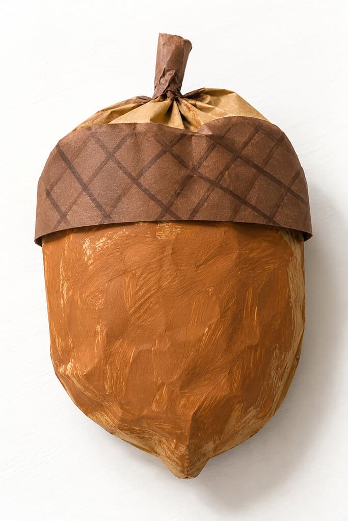 Acorn-shaped craft on a white surface with a painted brown body, a dark brown paper cap marked with a crosshatch pattern, and a crumpled paper stem at the top.