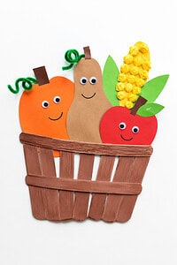 An orange circular pumpkin character, a tall tan pear character, and a red circular apple character, all with googly eyes and drawn smiles, alongside yellow crinkled tissue paper corn with green paper leaves, sitting inside a woven brown craft stick basket