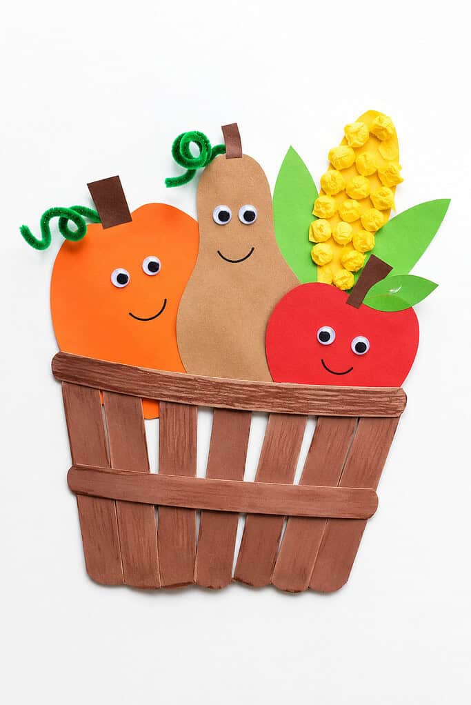 An orange circular pumpkin character, a tall tan pear character, and a red circular apple character, all with googly eyes and drawn smiles, alongside yellow crinkled tissue paper corn with green paper leaves, sitting inside a woven brown craft stick basket