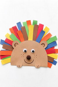Paper hedgehog craft with a tan face, two large white eyes with black pupils, a black nose, and a fan of rainbow-colored paper strip spikes