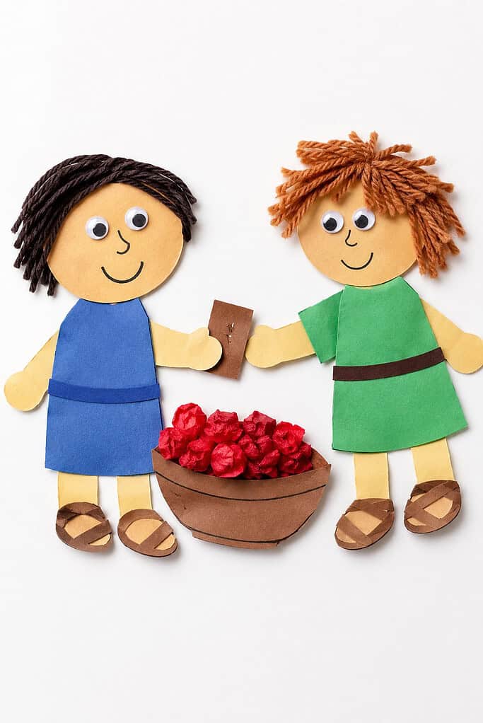 Two paper craft figures with yarn hair, one in a blue robe with black hair and one in a green robe with orange curly hair, standing beside a brown paper bowl filled with red crumpled paper, holding a small brown paper piece between their hands.