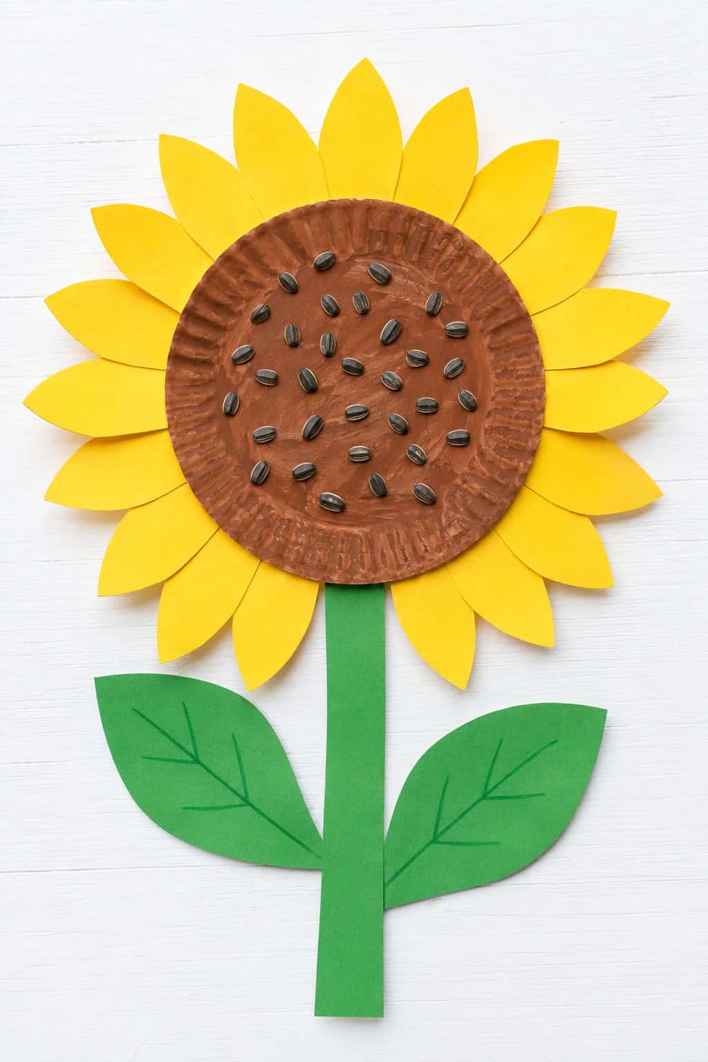 Paper plate sunflower craft with yellow paper petals, a brown painted center with sunflower seeds, a green paper stem, and two leaves on a white table.