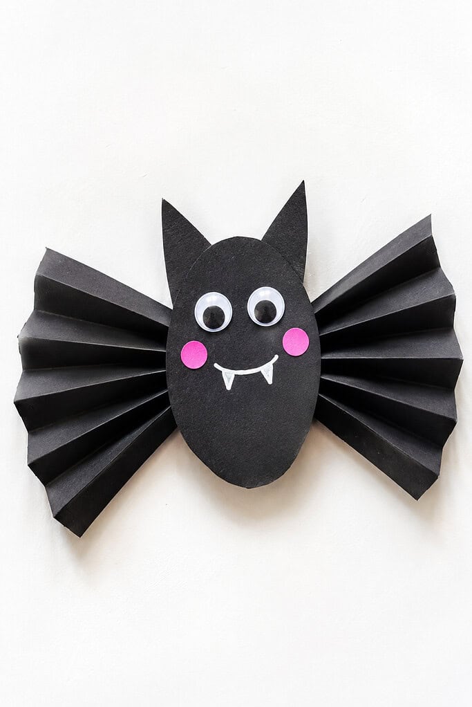 Black paper bat craft with accordion-folded wings, an oval body, pointed ears, googly eyes, pink cheeks, and a white smiling mouth on a bright white table.