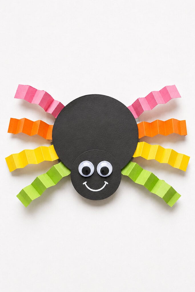 Black paper circle spider with googly eyes, a smile, and accordion-folded paper legs in pink, orange, yellow, and green on a white background