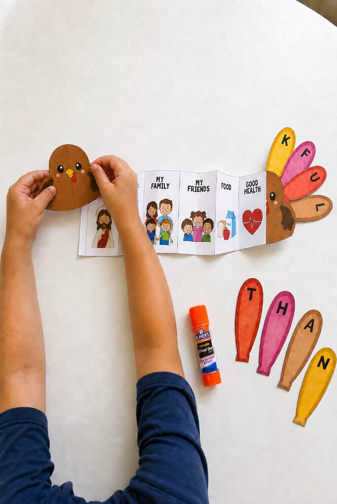 A child's hands gluing a small paper turkey onto the end of a folded accordion strip, with colorful lettered feathers laid out beside it on a white table