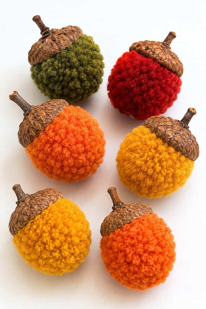 Six fuzzy, round acorn-shaped objects with brown textured caps and short stems are arranged on a white surface. Their soft yarn-like bodies are colored olive green, red, bright orange, golden yellow, and mustard yellow.