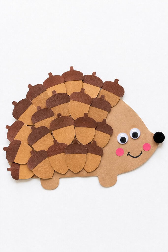 Paper acorn cutouts overlapping to form the spiky back of a hedgehog, with a tan paper face showing googly eyes, a black pom-pom nose, pink cheeks, and a smile