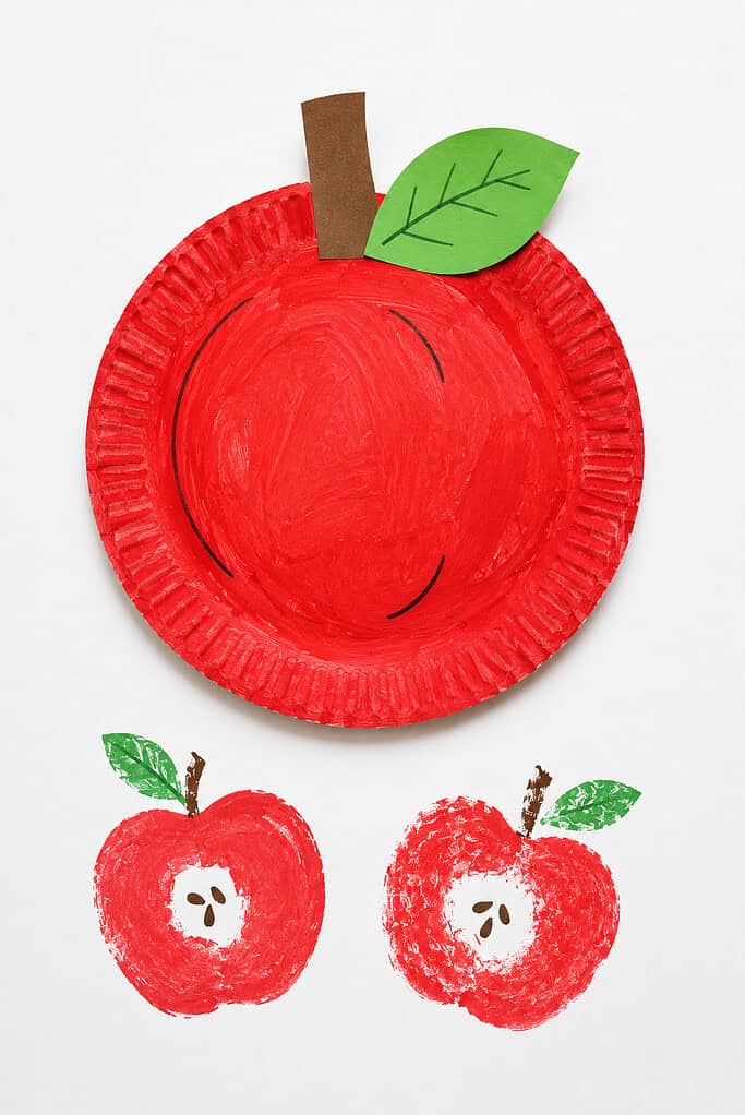 A red-painted paper plate with a brown paper stem and a green paper leaf with drawn veins at the top, with two red apple stamp prints below showing white centers with seeds, each with a green painted leaf and brown stem