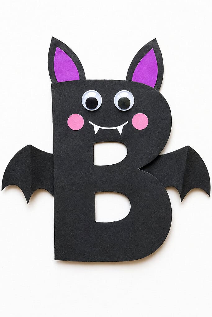 Large black paper letter B with side wings, pointed ears, purple inner ears, googly eyes, pink cheeks, and a white smiling mouth with two fangs on a white table.