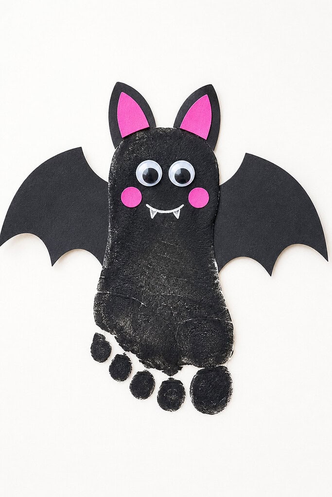 Black painted footprint with five visible toe prints, paper bat wings, pink inner ears and cheeks, googly eyes, a white smile, and two small fangs on a white background.