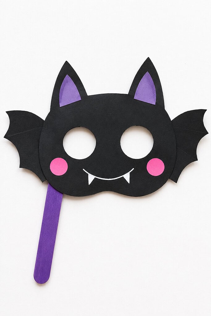 Black cardstock bat mask with circular eye holes, pointed ears, side wings, purple inner ears, pink cheeks, white fangs, and a purple craft-stick handle on a white table.