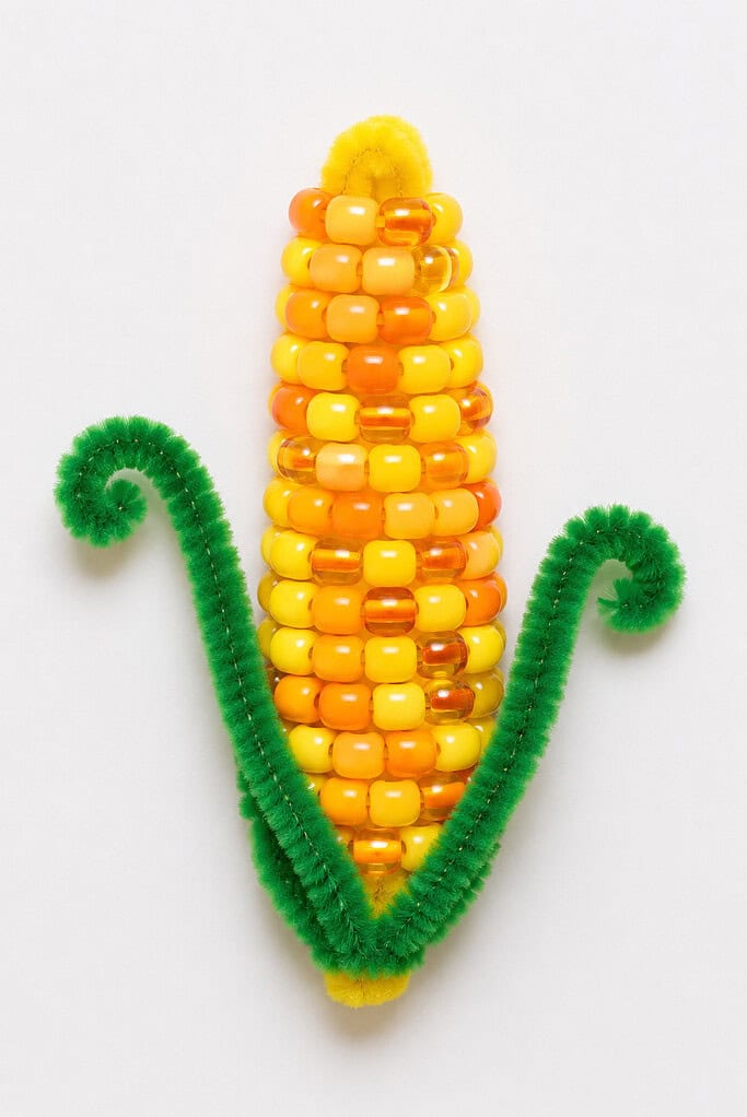 Yellow and orange plastic beads threaded onto a pipe cleaner in a corn cob shape, with two green fuzzy pipe cleaners curled on each side as husks, on a white background
