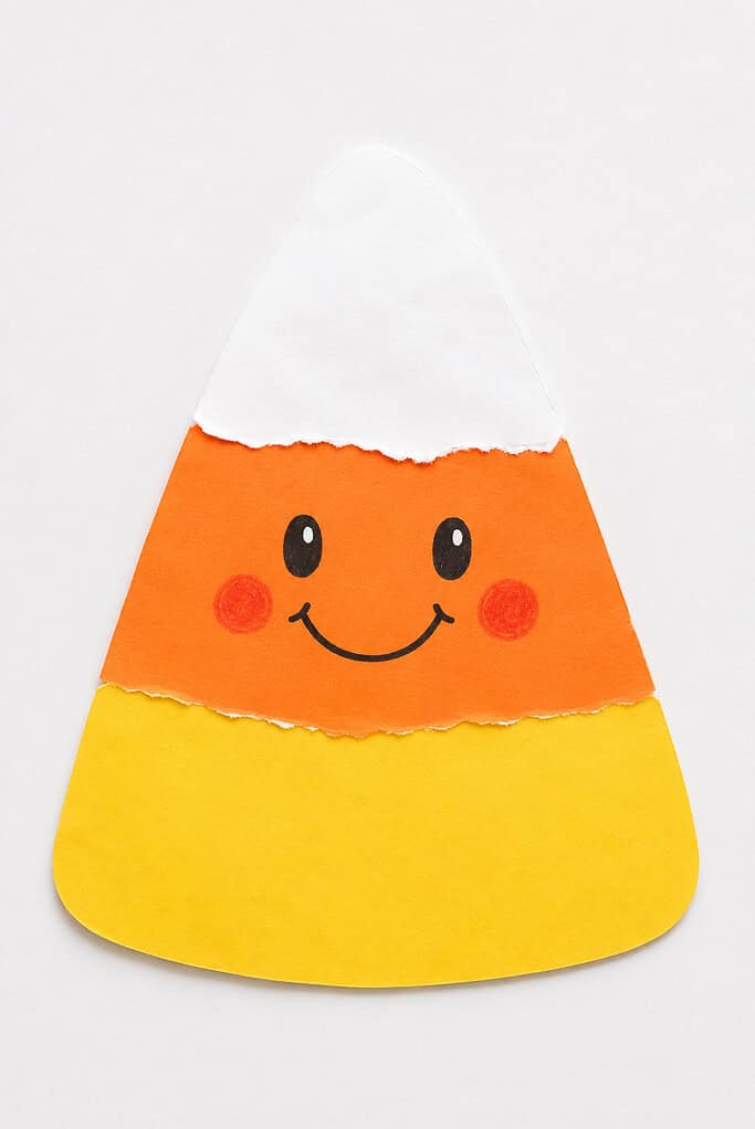 A large triangular shape made from three layers of torn paper — white on top, orange in the middle with drawn oval eyes, red pom-pom cheeks, and a black smile, and yellow at the bottom