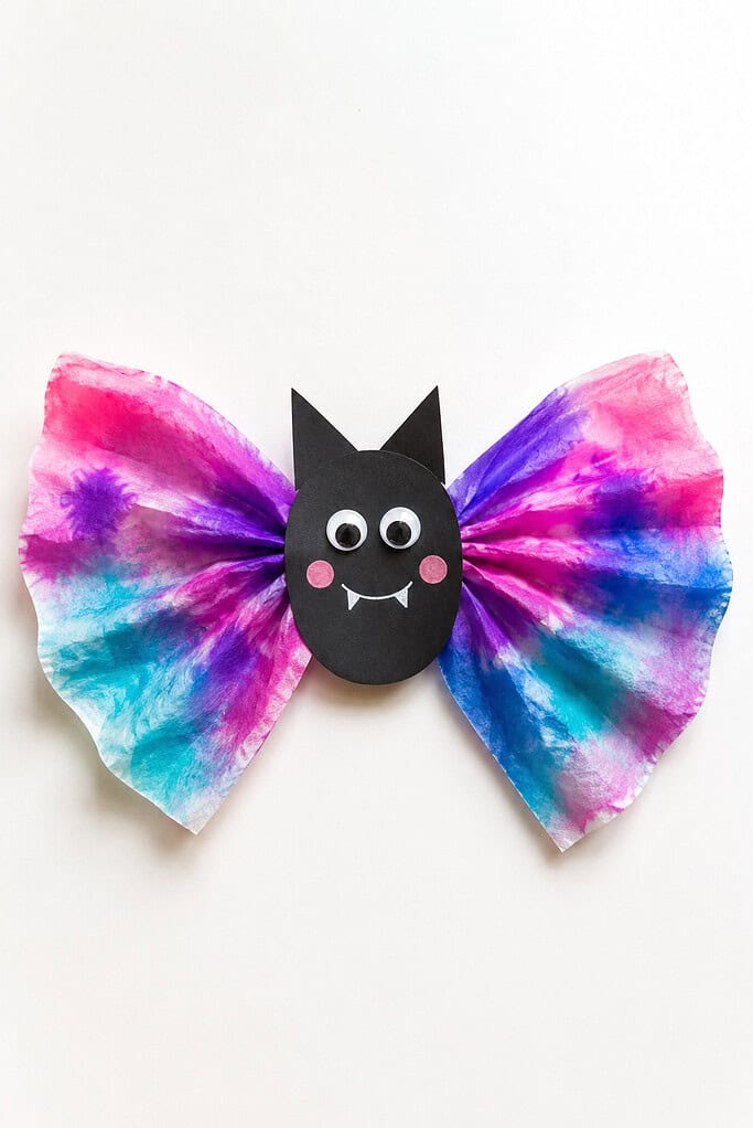 Black paper bat with pointed ears, googly eyes, pink cheeks, and a white smiling mouth attached to wide coffee filter wings in pink, purple, blue, and teal on a bright white table.