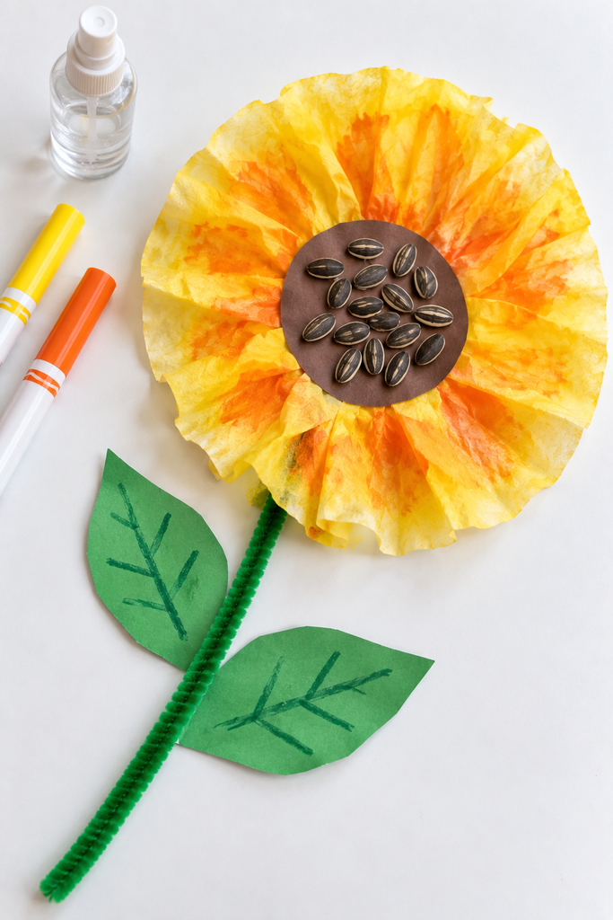 A coffee filter sunflower with ruffled yellow and orange petals in a tie-dyed look and a brown seeded center, the green pipe-cleaner stem running diagonally, with markers and a spray bottle at the edge of a white table.