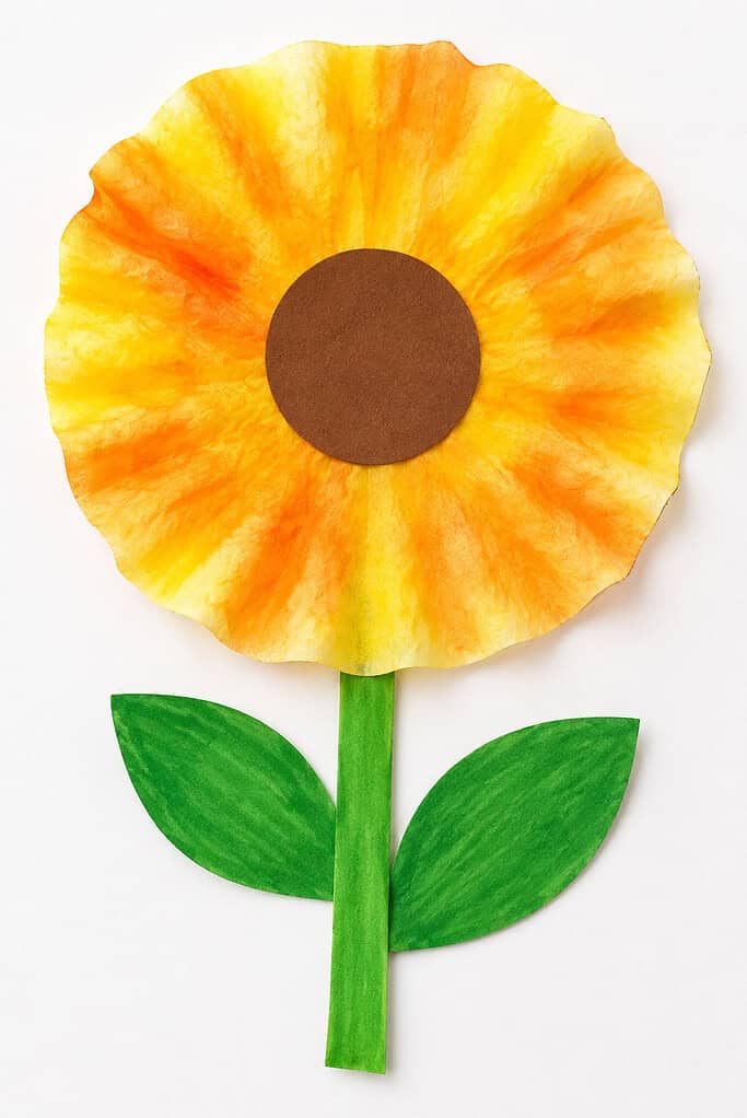 Yellow and orange coffee filter sunflower with a brown paper center, green stem, and two leaves on a white table.