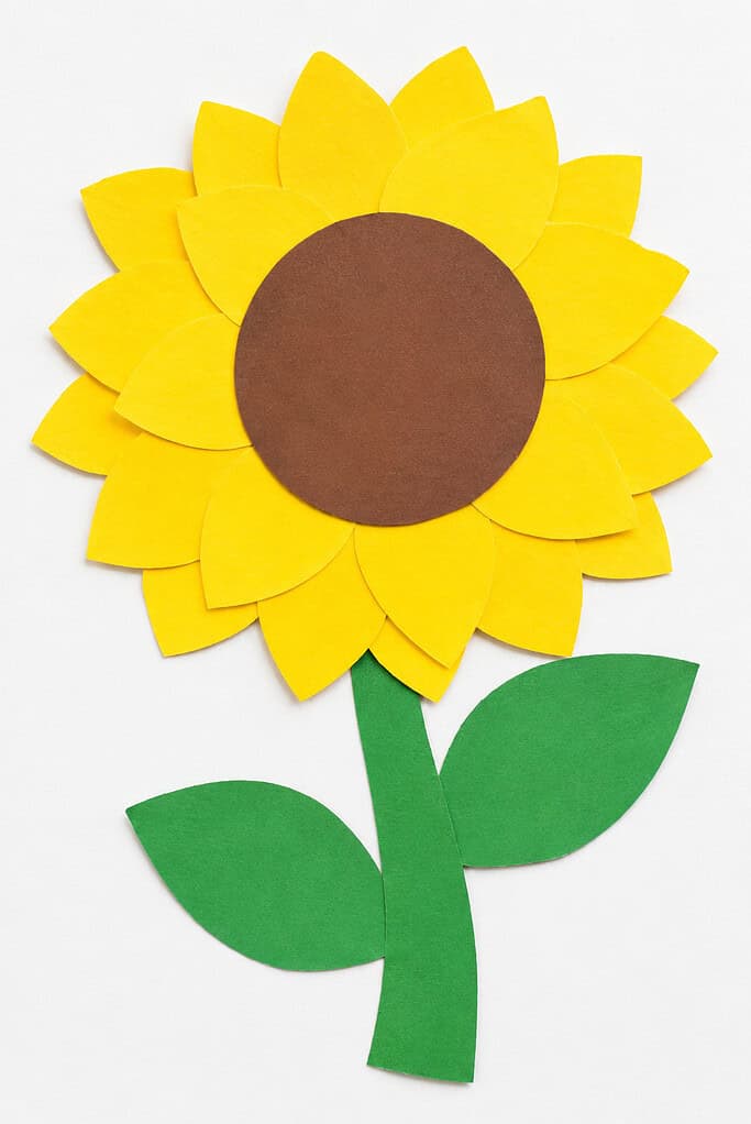 Flat lay of a construction paper sunflower with layered yellow petals, a large brown center, and a green stem with two green leaves on a bright white table.