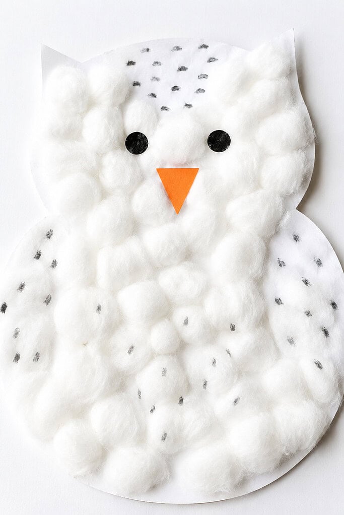 Owl body covered in white cotton balls with two small black dot eyes, an orange triangular paper beak, and gray marker speckles across the fluffy surface.