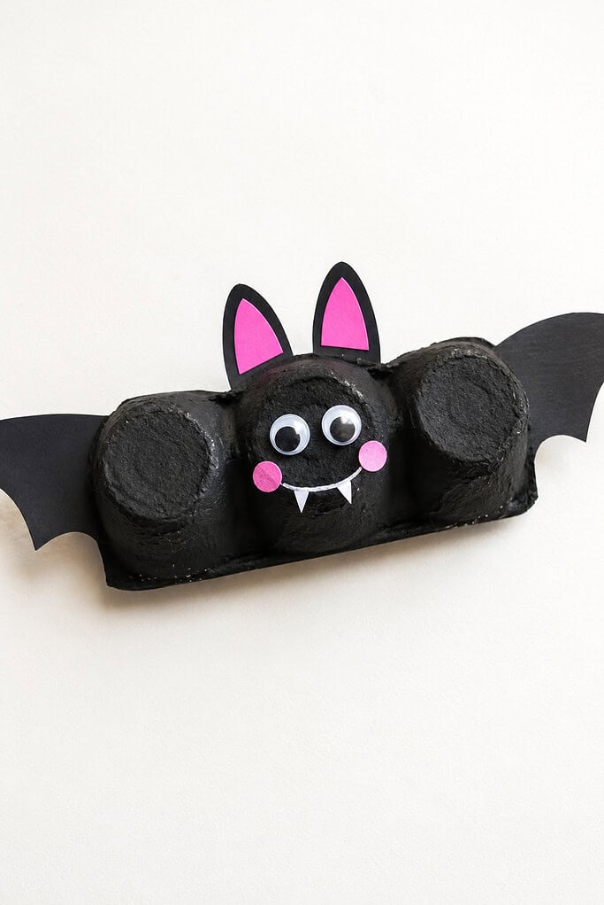 Black-painted three-cup egg carton with paper wings, pink inner ears and cheeks, googly eyes, a white smile, and two small fangs on a white table.