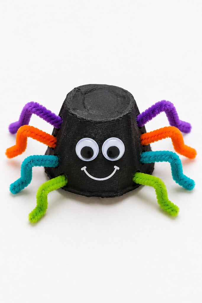Black-painted egg carton cup spider with googly eyes, a smile, and colorful pipe cleaner legs in purple, orange, teal, and green poked through the sides