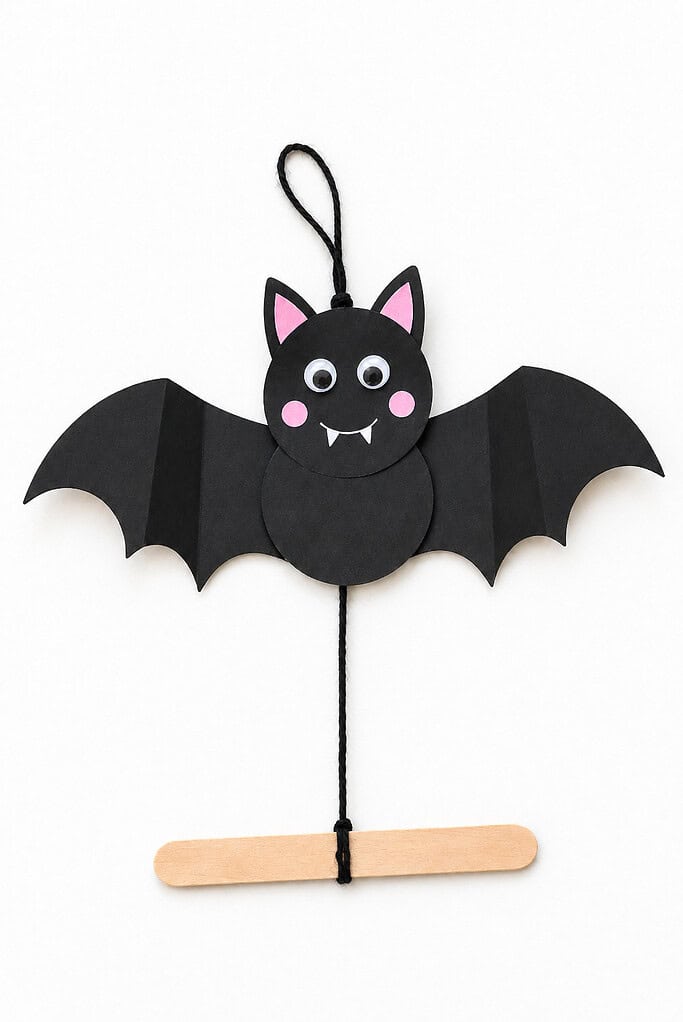 Black paper bat with wide folded wings, pink ears and cheeks, googly eyes, and white fangs, with black string tied to its head and a wooden craft stick below.