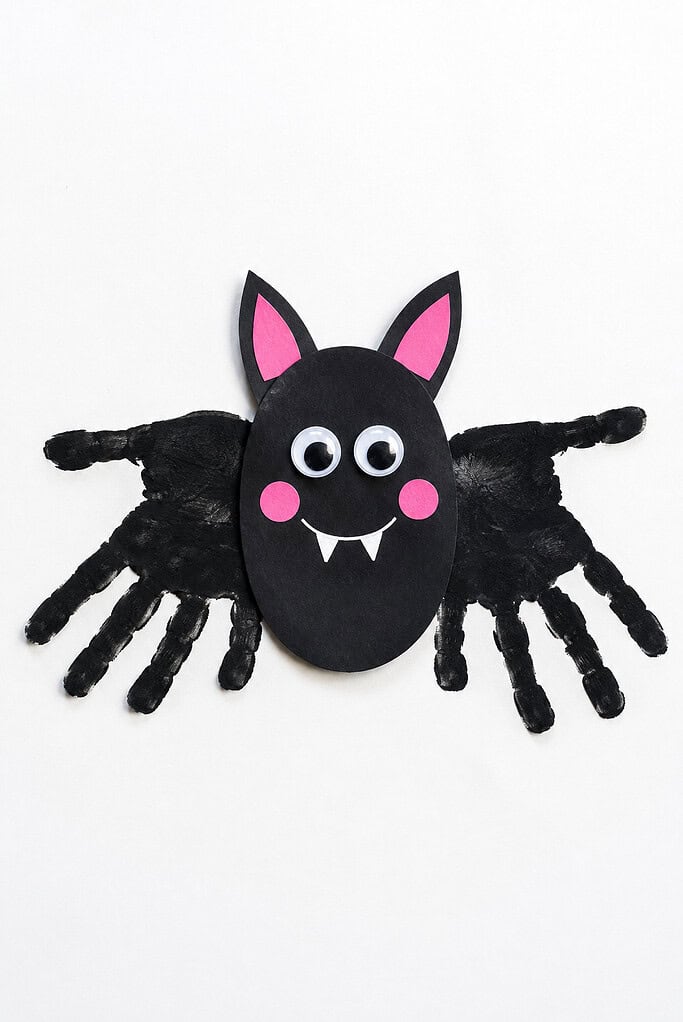 Black handprint wings beside an oval paper body with pink ears and cheeks, googly eyes, a white smile, and two small fangs on a white background.
