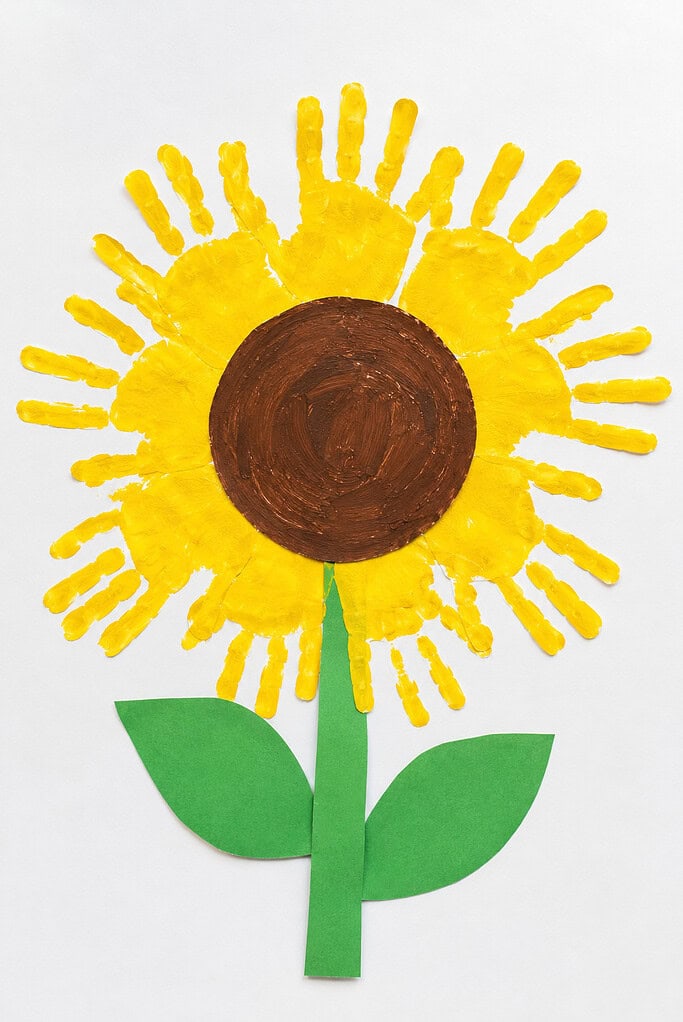 Yellow painted handprints arranged around a brown circular center with a green paper stem and two leaves on a white table.
