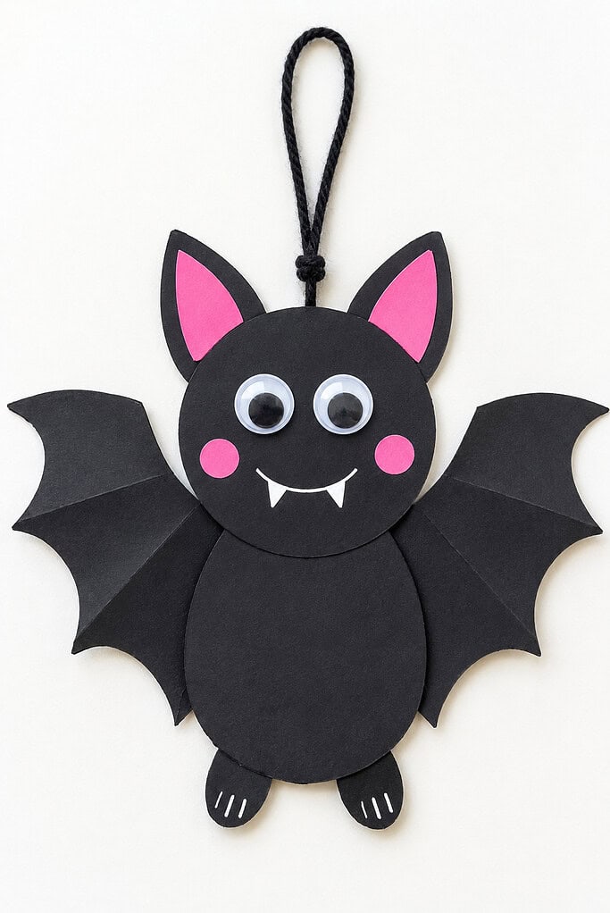 Black paper bat with a yarn hanging loop, wide folded wings, pink inner ears and cheeks, googly eyes, a white smile, two small fangs, and feet with white claw marks.