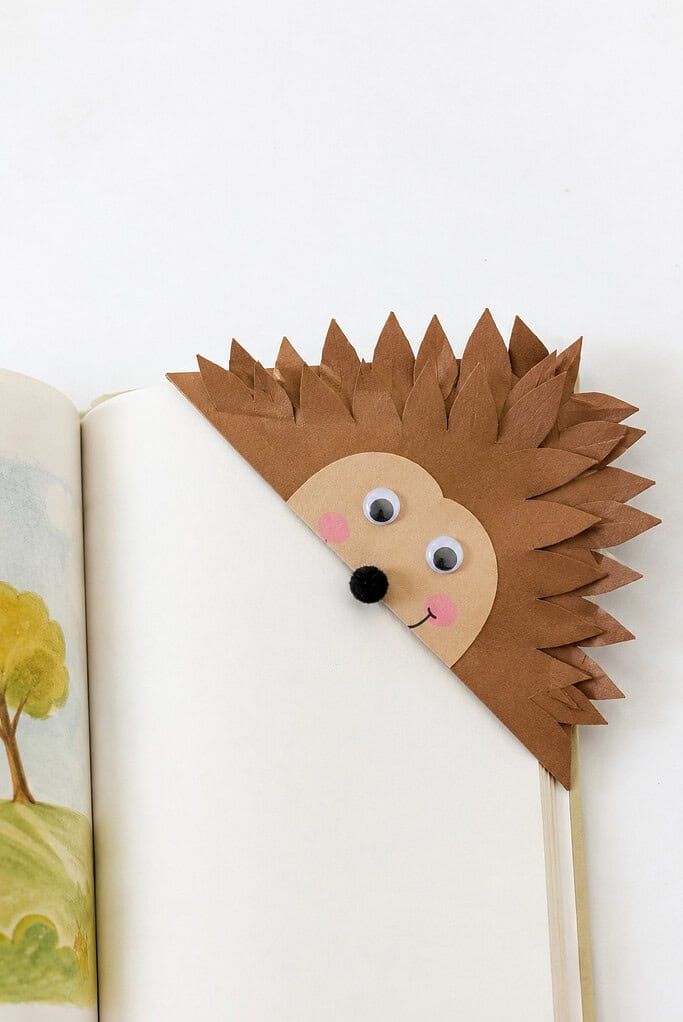 Brown paper corner bookmark shaped like a hedgehog face with pointed spiky quills, googly eyes, pink cheeks, a black pom-pom nose, and a smile, slipped onto the corner of a book page