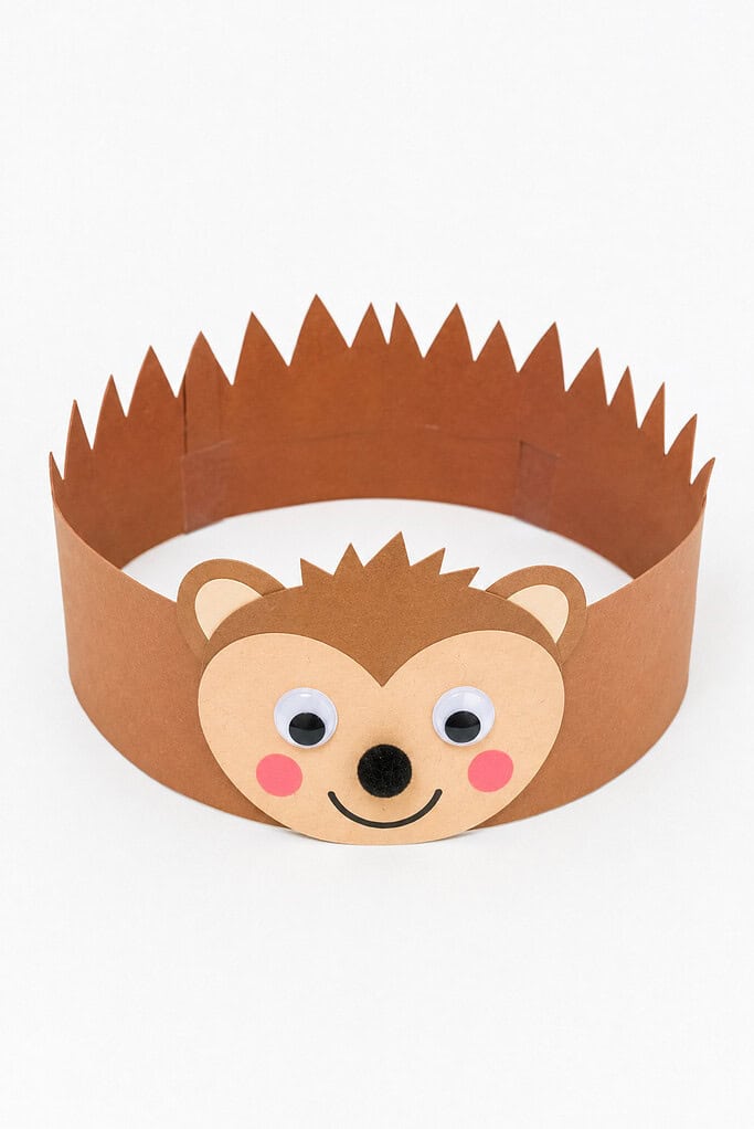 Wearable paper headband with brown triangular spikes around the top and a hedgehog face on the front showing rounded ears, googly eyes, a black pom-pom nose, pink cheeks, and a smile