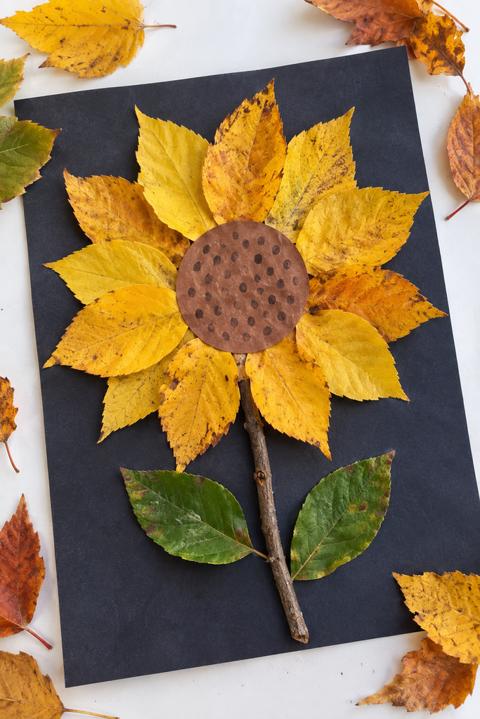 A sunflower craft made from real leaves on dark paper, with golden autumn leaves as petals around a brown crosshatched center, a real twig stem, and green leaves as foliage, surrounded by scattered autumn leaves.