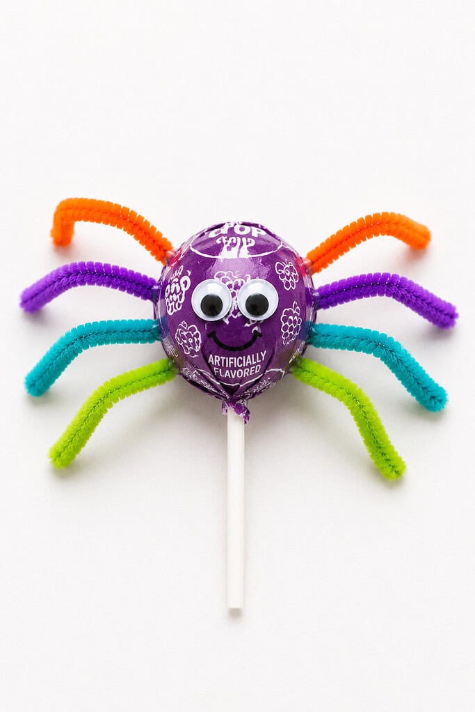 Purple wrapped lollipop spider with a drawn googly-eyed smiling face and colorful pipe cleaner legs in orange, purple, teal, and green, on a stick