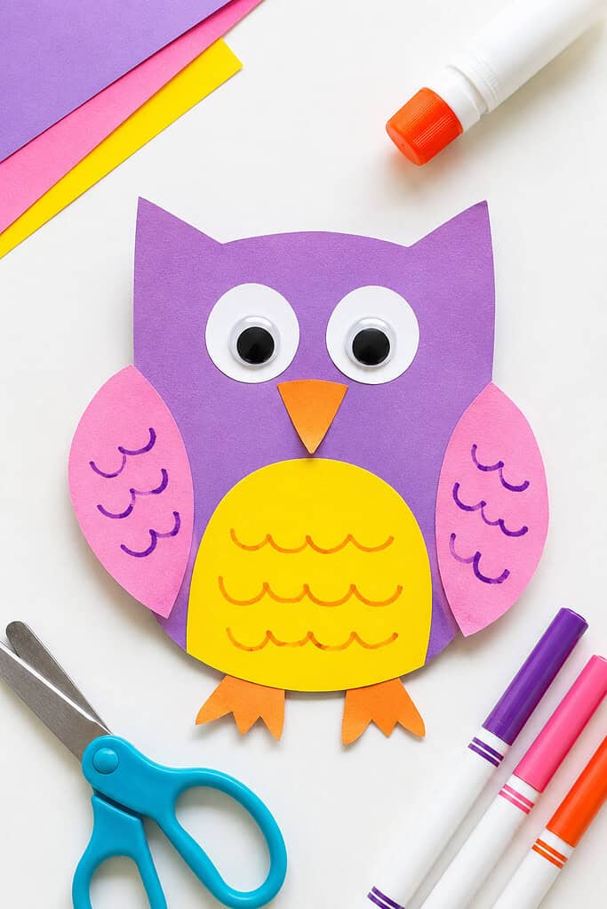 Purple felt owl craft with pink wings featuring yellow scalloped edges, a yellow belly, large blue and white eyes, and an orange triangular beak, displayed with scissors and paper scraps nearby.