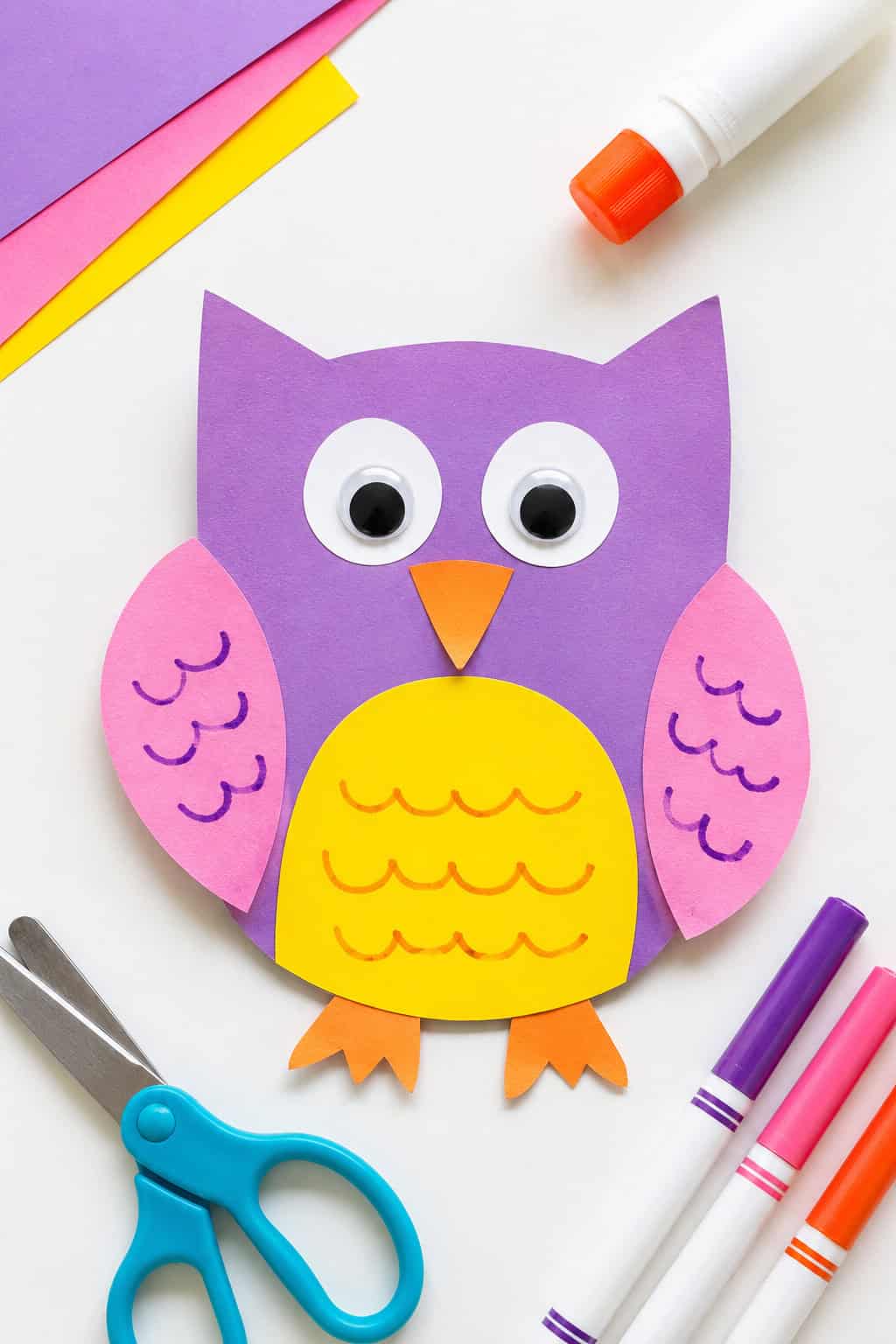 Purple felt owl craft with pink wings featuring yellow scalloped edges, a yellow belly, large blue and white eyes, and an orange triangular beak, displayed with scissors and paper scraps nearby.
