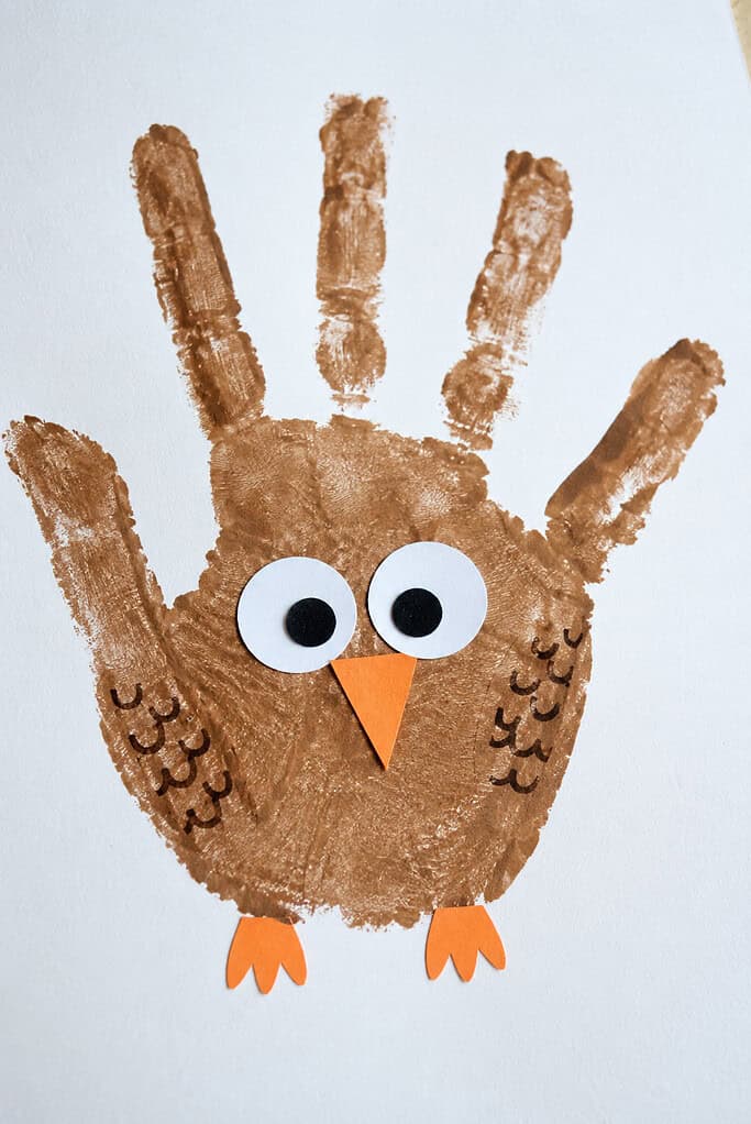 Brown painted handprint on paper shaped like an owl, with fingers as feathers, two large white googly eyes with black pupils, an orange triangular beak, and orange feet.