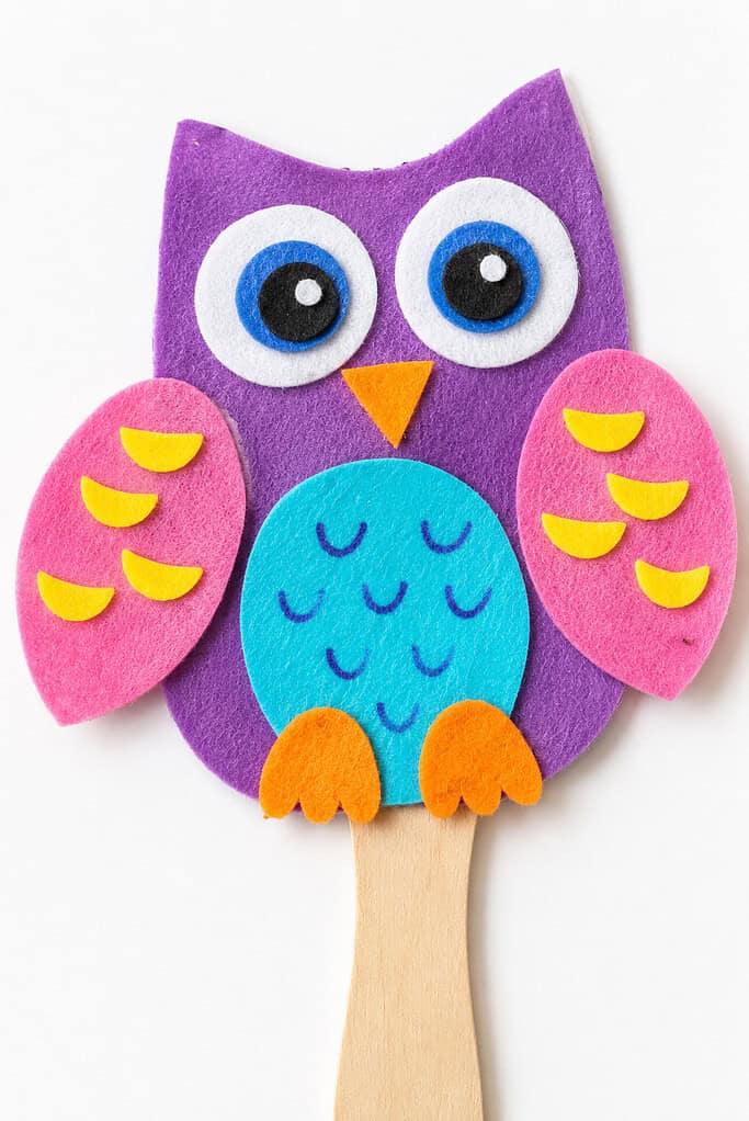 Felt owl puppet on a wooden stick, with a purple face, large blue and white eyes, an orange triangular beak, pink wings with yellow crescent shapes, a teal belly with stitched arch patterns, and orange feet.
