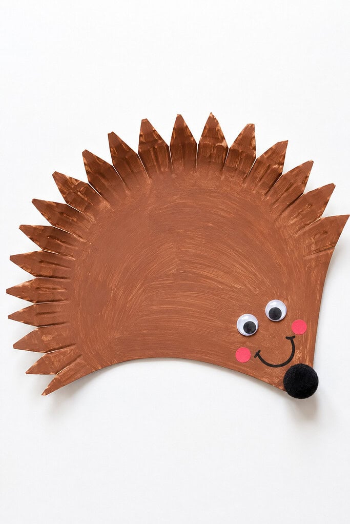 Paper plate painted brown with the edge cut into spiky points, googly eyes, pink cheeks, a black pom-pom nose, and a smile painted on