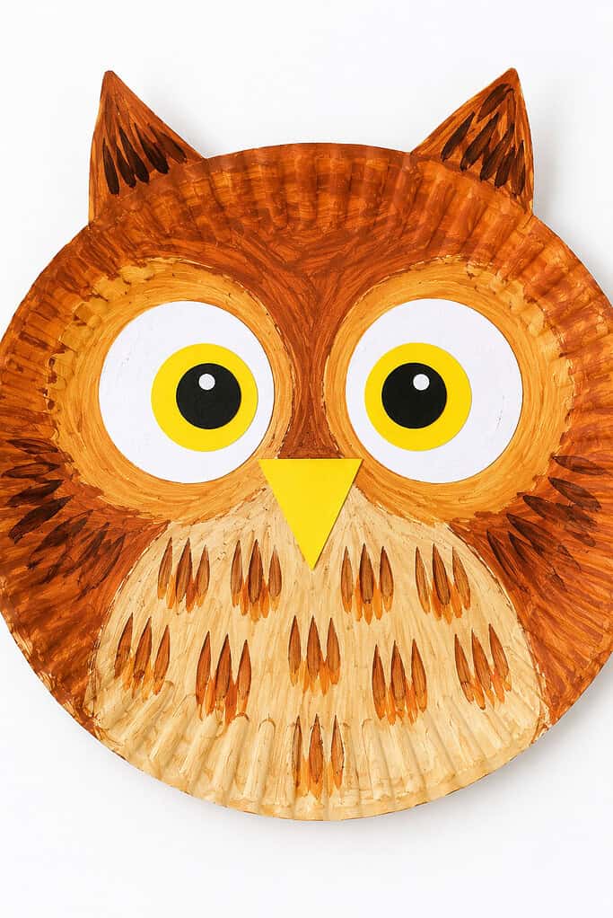 Paper plate painted orange and brown with feather-like brush strokes, pointed ear tufts, large white and yellow eyes with black pupils, and a yellow triangular beak.