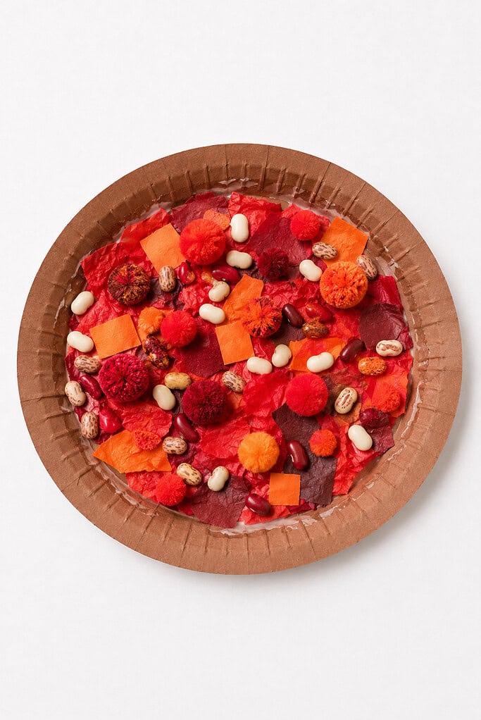 A brown paper plate filled with red paper or paint texture topped with orange paper squares, white beans, dried beans, and small red and orange pom-poms.