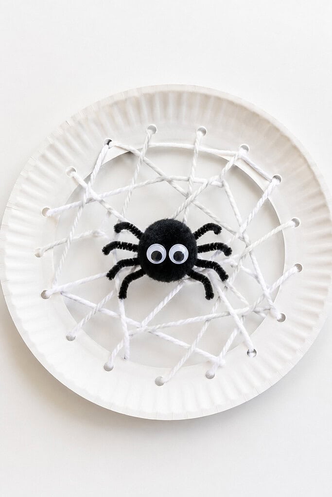 White paper plate with yarn woven into a web pattern and a black pom-pom spider with googly eyes in the center