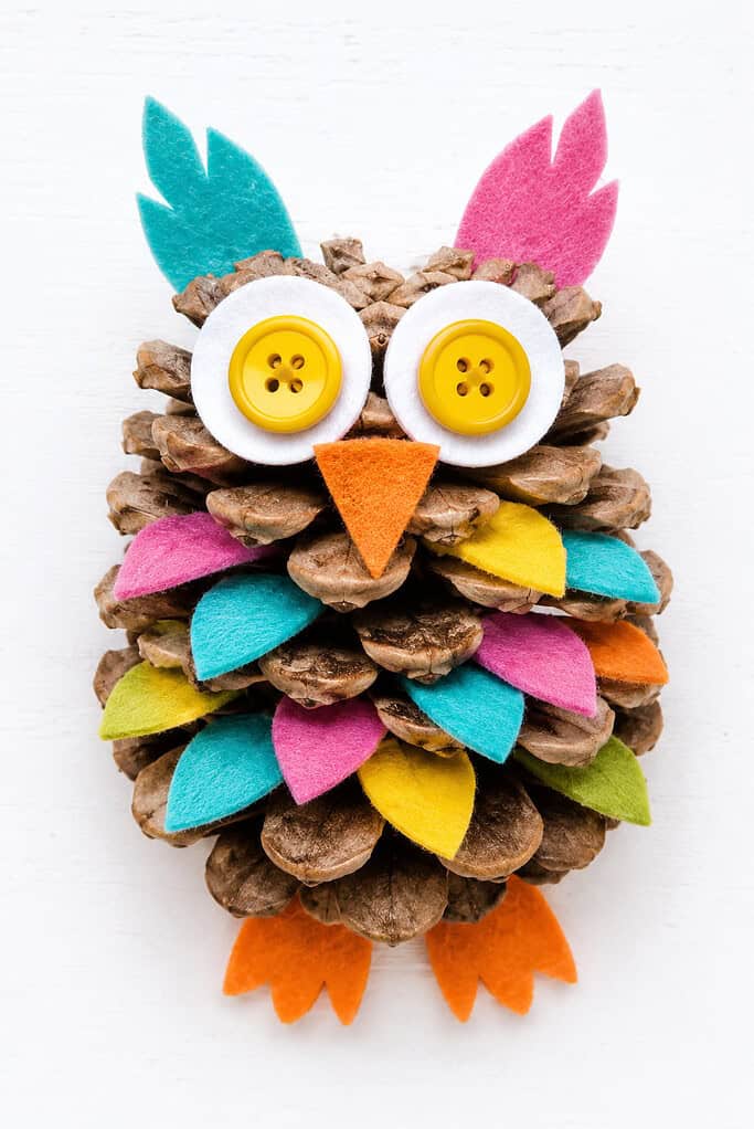 Pinecone decorated as an owl with yellow button eyes on white felt circles, an orange felt triangular beak, and colorful felt feather pieces in teal, orange, yellow, and green tucked between the pinecone scales.
