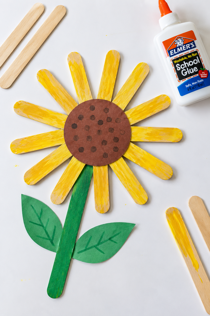 A popsicle stick sunflower with yellow-painted craft sticks arranged like spokes around a brown seeded center, with a green stick stem and paper leaves, and a few loose craft sticks and glue at the edge of a white table.