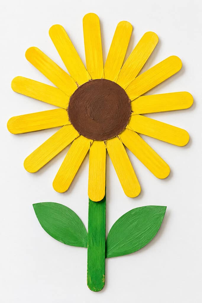 Flat lay of a sunflower craft made with yellow painted popsicle sticks, a round brown center, a green popsicle stick stem, and two green leaves on a white table.