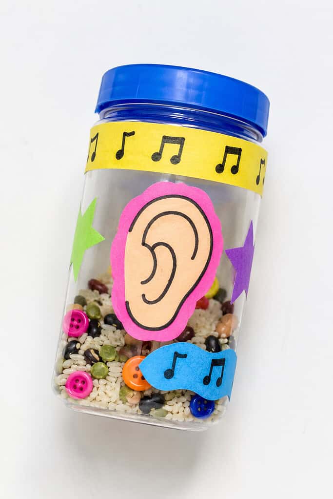 Clear plastic jar with a blue lid, filled with rice, beans, and colorful buttons, and decorated with paper musical notes, stars, and an ear cutout.