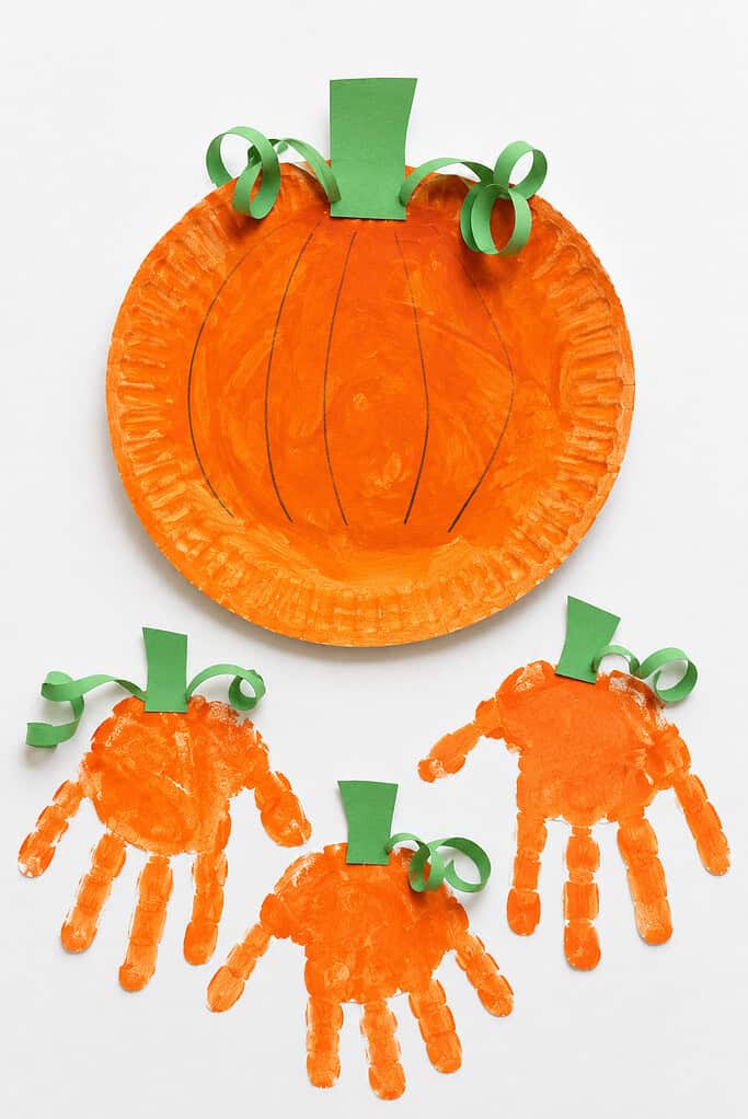 A large orange-painted paper plate with black vertical lines and a green paper stem and curly tendrils, alongside three smaller orange handprint shapes each with green paper stems and curly tendrils, on a white background